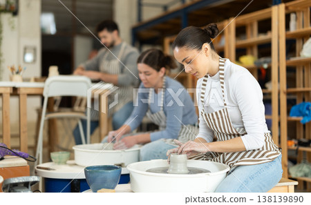 Girl craftspersons busy molding clay products on pottery wheel Girl craftspersons busy molding clay products on pottery wheel 138139890