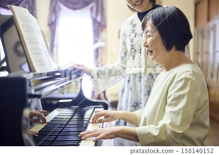 A senior woman plays the piano with a smile, watched over by a female instructor. A senior woman plays the piano with a smile, watched over by a female instructor. 138140152