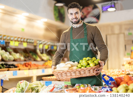Man employees in uniform carries wicker basket with various vegetables and fruits in grocery shop Man employees in uniform carries wicker basket with various vegetables and fruits in grocery shop 138140240