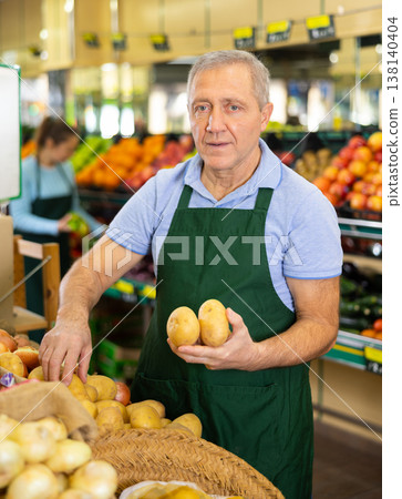 Skilled elderly male seller demonstrating local potatoes in shop 138140404