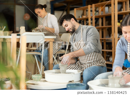 Man in ceramics workshop with pottery wheel and various clay vessels 138140425