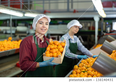 Glad positive female employee in colored uniforms hold a box of fresh ripe tangerines in their hands on citrus sorting line at warehouse. Glad positive female employee in colored uniforms hold a box of fresh ripe tangerines in their hands on citrus sorting line at warehouse. 138140431