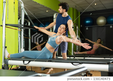 Woman doing abdominal exercises with the help of a male trainer and a reformer Woman doing abdominal exercises with the help of a male trainer and a reformer 138140473