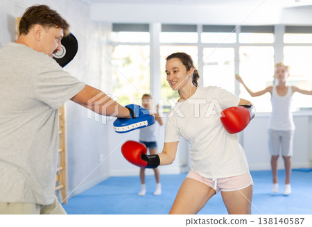 Positive woman in boxing gloves practicing punches on mitts with husband Positive woman in boxing gloves practicing punches on mitts with husband 138140587