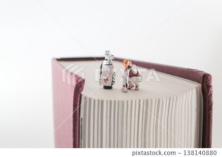 Miniature Golfer Kneels Beside Bag on Oversized Book 138140880