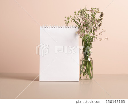 The planner's blank clean notepad stands on the table in vase with delicate green flowers. Mock up for text The planner's blank clean notepad stands on the table in vase with delicate green flowers. Mock up for text 138142163