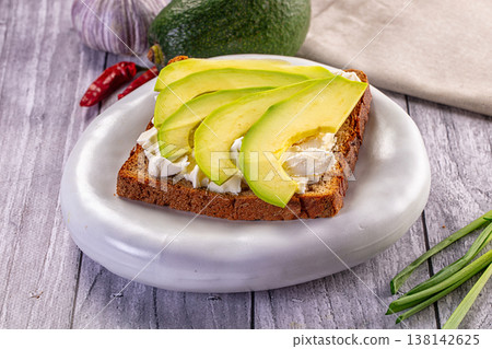 Avocado toast with soft cheese on dark rye bread. A healthy breakfast sandwich on a white plate over a rustic wooden table. 138142625