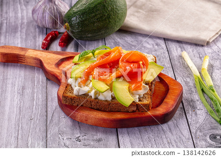 Gourmet toast with smoked salmon, avocado, and soft cheese on a rustic wooden table with fresh ingredients. 138142626