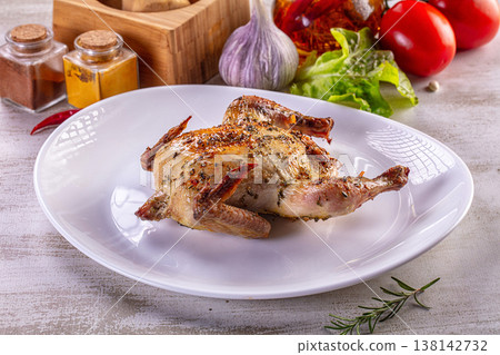 Roasted quail with herbs on a white plate surrounded by fresh vegetables and spices. 138142732