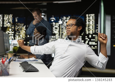 Tired engineer stretching at office desk while working late on computer for tech company. Exhausted man relaxing muscles, feeling fatigue while doing machine learning coding on PC 138145283