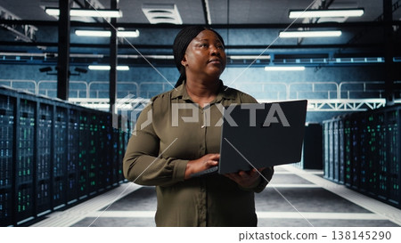 Admin walks between server farm rows, using laptop to upgrade rigs for increase in clients workload capacity. Data center worker configures hardware with notebook, improving processing speed, camera A Admin walks between server farm rows, using laptop to upgrade rigs for increase in clients workload capacity. Data center worker configures hardware with notebook, improving processing speed, camera A 138145290