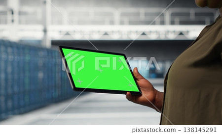IT professional in data center using green screen tablet to ensure error free supercomputers networks. African american woman uses mockup device to monitor server hub gear parts for failures, camera B IT professional in data center using green screen tablet to ensure error free supercomputers networks. African american woman uses mockup device to monitor server hub gear parts for failures, camera B 138145291