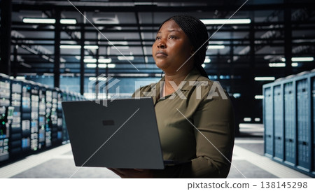 Technician using laptop in high tech data center, integrating new technologies and systems. IT expert using notebook to oversee server farm rigs, ensuring compliance with industry standards, camera A 138145298