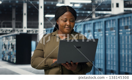 Programmer in data center using laptop to upgrade systems for increased stability. IT professional in server farm configuring infrastructure with notebook for optimum performance, camera B 138145308