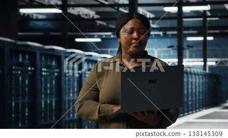 Data center system administrator uses laptop to develop disaster recovery plans, minimizing downtime. IT specialist walking in server farm, enhancing data protection protocols using notebook, camera B Data center system administrator uses laptop to develop disaster recovery plans, minimizing downtime. IT specialist walking in server farm, enhancing data protection protocols using notebook, camera B 138145309
