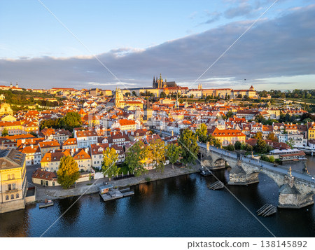 In the early morning, sunlight washes over the rooftops of Prague, highlighting the iconic Charles Bridge and the majestic Prague Castle in the background, along the Vltava River. 138145892