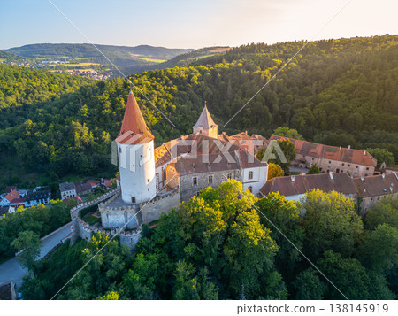 Krivoklat Castle stands majestically in central Bohemia, surrounded by lush green forests. The warm hues of sunset illuminate the medieval architecture, revealing its historical beauty. 138145919