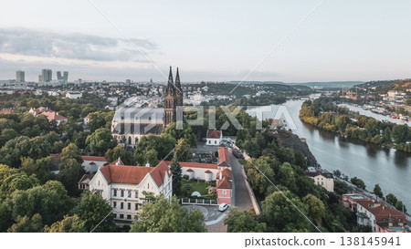 Morning sunlight bathes the majestic Vysehrad Basilica of St. Peter and St. Paul in Prague. The serene landscape features lush greenery, historic architecture, and the flowing Vltava River. 138145941