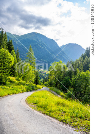 A road curves through lush green trees and grass in the Alpine region. Mountains rise in the distance under a partly cloudy sky. It is a sunny day in summer. A road curves through lush green trees and grass in the Alpine region. Mountains rise in the distance under a partly cloudy sky. It is a sunny day in summer. 138145956