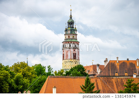 The castle tower stands tall in Cesky Krumlov surrounded by trees. The town features traditional buildings with tiled roofs under a cloudy sky. Visitors explore the streets below in summer. 138145974