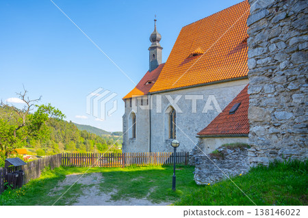 Visitors walk near Holy Trinity Church in Czechia during a sunny day. The church stands with a stone wall and red roof. Green hills surround the area, creating a scenic view for all. 138146022