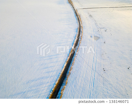 An aerial view of an asphalt road winding through a snowy field. The scene captures the contrast between the dark road and bright white snow. The photo shows winter conditions. 138146071