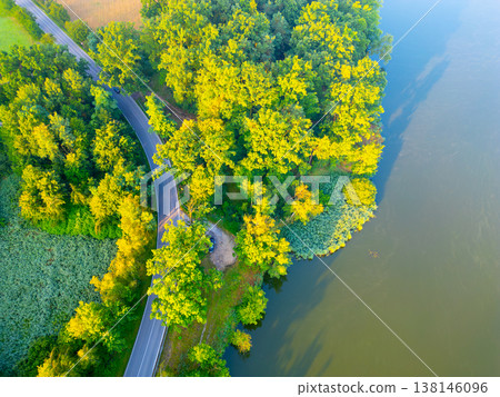 A winding road runs alongside a body of water. Trees surround the area with a mix of green foliage. The scene is captured from an aerial view during daylight. 138146096