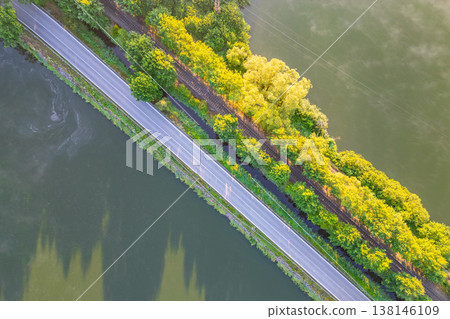 A view shows a road and railroad running next to a body of water. Trees line the edges, creating a green border. This scene captures the intersection of land and water. A view shows a road and railroad running next to a body of water. Trees line the edges, creating a green border. This scene captures the intersection of land and water. 138146109