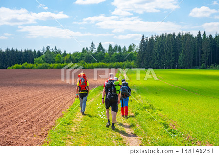Four people walk on a path through agricultural land in Nova Paka, Czechia. Red clay soil and green fields are visible under a bright blue sky with clouds. 138146231