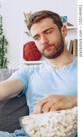 Man sitting on a couch eating popcorn while watching basketball in a living room 138146901