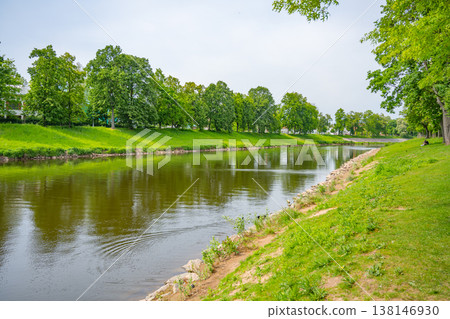 Jirsakovy sady in Hradec Kralova shows green trees along the riverside. People are walking and sitting on the grass. The Elbe and Orlice rivers meet in this area. Jirsakovy sady in Hradec Kralova shows green trees along the riverside. People are walking and sitting on the grass. The Elbe and Orlice rivers meet in this area. 138146930