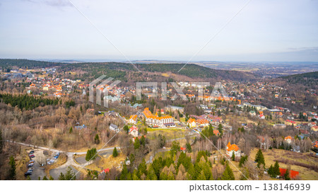 A wide view of a small town surrounded by trees and hills. Several buildings are visible, with roads and cars parked nearby. The scene shows a clear sky above. A wide view of a small town surrounded by trees and hills. Several buildings are visible, with roads and cars parked nearby. The scene shows a clear sky above. 138146939
