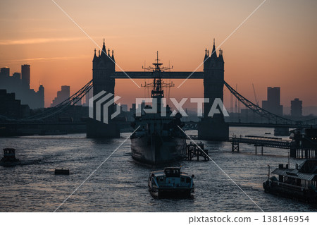 A dawn scene on the River Thames shows HMS Belfast and the silhouette of Tower Bridge. Boats move along the river as the sun rises, casting a warm glow over London. A dawn scene on the River Thames shows HMS Belfast and the silhouette of Tower Bridge. Boats move along the river as the sun rises, casting a warm glow over London. 138146954