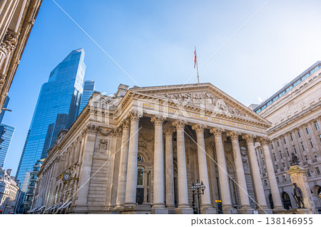 The Royal Exchange in London stands among modern structures on a clear day. People walk nearby, and the British flag flies high above the historic site. 138146955