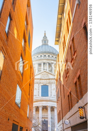 A narrow street leads to St Pauls Cathedral in London. The tall structure is visible between the red brick buildings. The sunlight shines on the historic architecture. A narrow street leads to St Pauls Cathedral in London. The tall structure is visible between the red brick buildings. The sunlight shines on the historic architecture. 138146956