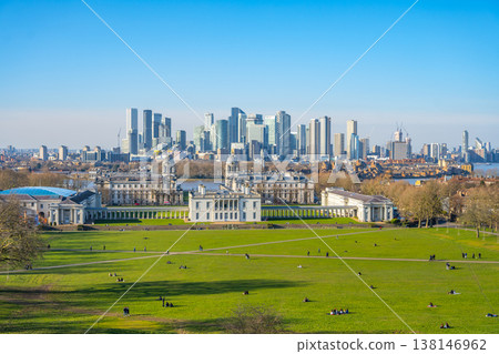 Visitors enjoy a sunny day at Greenwich Park, surrounded by lush green grass and historic buildings. The skyline of modern London towers in the background, showcasing the city's blend of old and new. 138146962