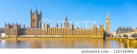 Houses of Parliament and Big Ben sit next to the River Thames. The sun shines brightly on this famous London landmark. People enjoy the view from the riverbank. Houses of Parliament and Big Ben sit next to the River Thames. The sun shines brightly on this famous London landmark. People enjoy the view from the riverbank. 138146964