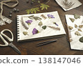 Creating botanical herbarium with pressed purple flowers and dried leaves in sketchbook. Creative workspace on wooden table with scissors, tweezers and crafting tools for DIY nature art project. 138147046