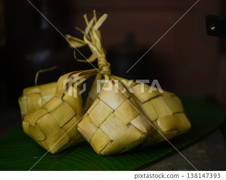 Traditional ketupat dumplings wrapped in woven palm leaves on a green banana leaf in Indonesia with warm tones and soft lighting. 138147393