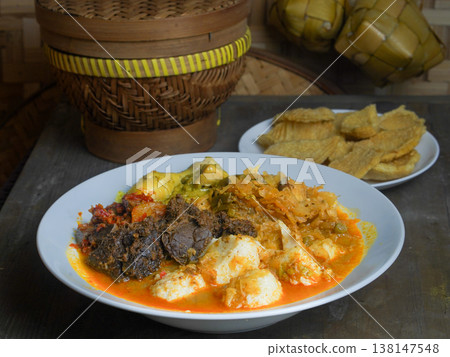 Traditional Indonesian ketupat sayur dish served on a white plate with a wooden container and food items in the background on a rustic table. Traditional Indonesian ketupat sayur dish served on a white plate with a wooden container and food items in the background on a rustic table. 138147548