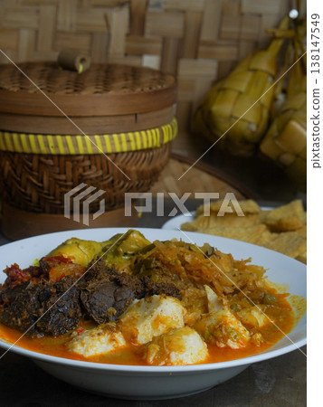 Traditional Indonesian ketupat sayur dish served on a white plate with a wooden container and food items in the background on a rustic table. Traditional Indonesian ketupat sayur dish served on a white plate with a wooden container and food items in the background on a rustic table. 138147549