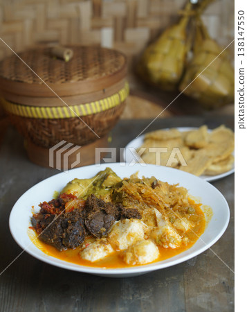 Traditional Indonesian ketupat sayur dish served on a white plate with a wooden container and food items in the background on a rustic table. Traditional Indonesian ketupat sayur dish served on a white plate with a wooden container and food items in the background on a rustic table. 138147550
