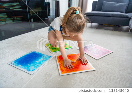 Little girl kneeling on floor pressing colorful sensory gel mats with hands during play activity. Sensory stimulation, autism support and occupational therapy development concept. Little girl kneeling on floor pressing colorful sensory gel mats with hands during play activity. Sensory stimulation, autism support and occupational therapy development concept. 138148640