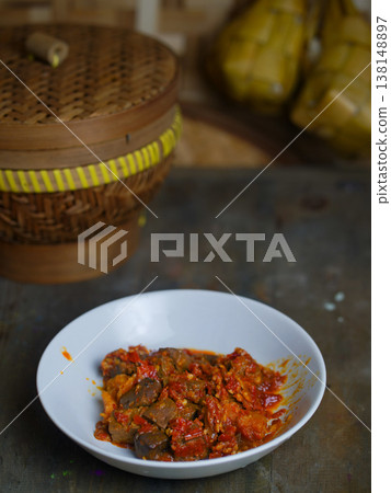 A plate of sambal goreng ati on a rustic table with a wicker steamer basket in the background, set against a blurred Indonesian kitchen backdrop with warm tones. A plate of sambal goreng ati on a rustic table with a wicker steamer basket in the background, set against a blurred Indonesian kitchen backdrop with warm tones. 138148897