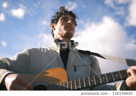 The wild and free spirit of the guitarist. A young guy plays the guitar against a background of blue sky and white clouds, a view from below. An inspired musician composes a new melody on the guitar The wild and free spirit of the guitarist. A young guy plays the guitar against a background of blue sky and white clouds, a view from below. An inspired musician composes a new melody on the guitar 138149685