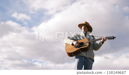 A smiling guitarist in a cowboy hat against a background of blue sky and white clouds. A single happy man is playing the guitar. The spirit of freedom A smiling guitarist in a cowboy hat against a background of blue sky and white clouds. A single happy man is playing the guitar. The spirit of freedom 138149687