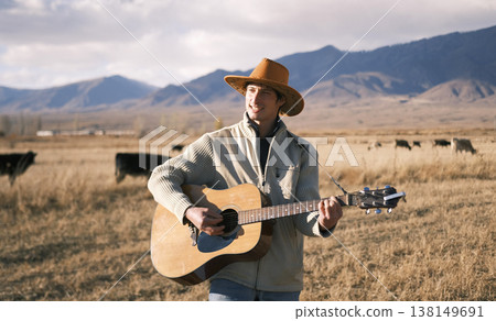 A handsome man in a cowboy hat sings songs with a guitar. A musician plays the guitar in a farmer's field. The sky is overcast, and mountains and a herd of cows A handsome man in a cowboy hat sings songs with a guitar. A musician plays the guitar in a farmer's field. The sky is overcast, and mountains and a herd of cows 138149691