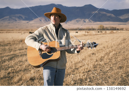 A drunken cowboy sings soulful songs with a guitar in a dry farmer's field. A man in a cowboy hat is playing guitar in the mountains. The guitarist gives a concert to his cows in the field A drunken cowboy sings soulful songs with a guitar in a dry farmer's field. A man in a cowboy hat is playing guitar in the mountains. The guitarist gives a concert to his cows in the field 138149692