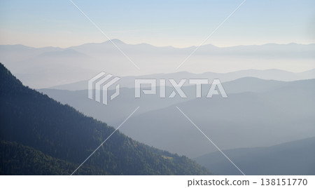 Misty morning in mountains with layers of forested hills fading into distance. Rising sun casts golden light on green slope, creating serene and peaceful landscape. Misty morning in mountains with layers of forested hills fading into distance. Rising sun casts golden light on green slope, creating serene and peaceful landscape. 138151770