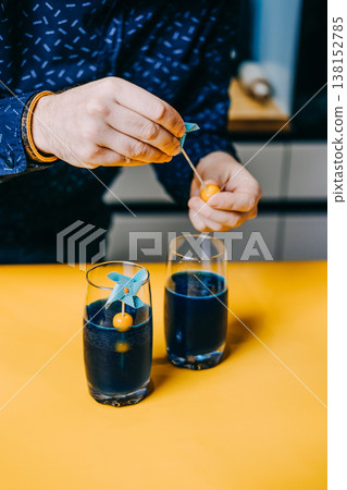 Hands assemble garnish with berry and pinwheel over glasses of blue liquid on yellow surface. blue spirulina mocktails, natural blue beverages, phycocyanin drink recipes, vibrant blue elixirs 138152785